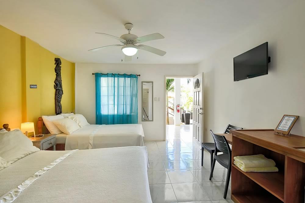 Bedroom of a gest house with two double beds, table and drawers and open door with view of the outside